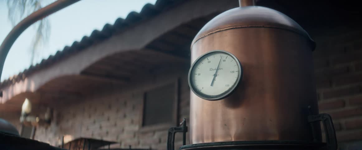 A closeup shot of the top section of a round copper distillation tank a white and black circular temperature gauge is attached to the tank side a portion of an overhanging roof and a stone wall are in the background the scene is lit by sunlight
