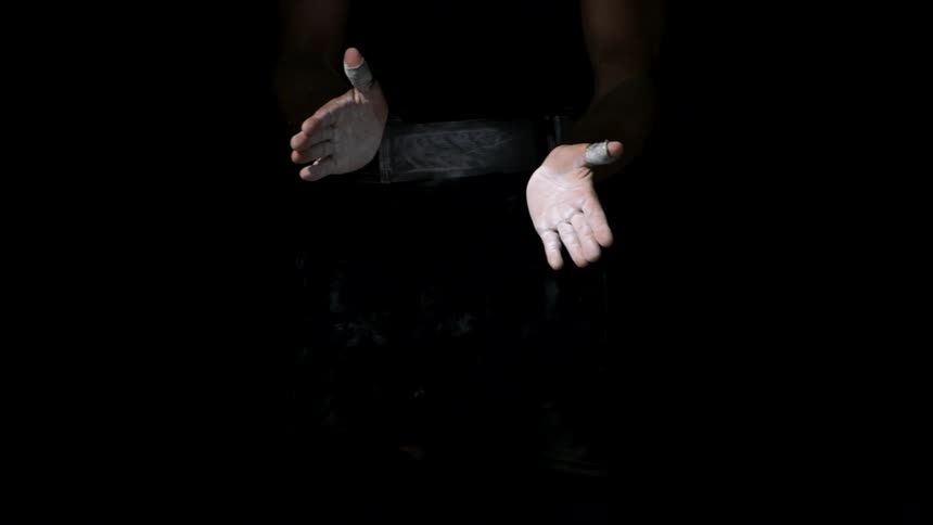 A pair of chalk covered hands clap in a dark scene sending a burst of white powder into the air as muscular forearms emerge from the shadows