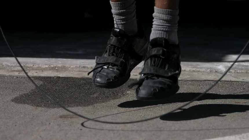 The video is a lowangle closeup shot of a persons legs jump roping on pavement outdoors they are wearing black training or cycling shoes and light grey socks that extend high up their calves