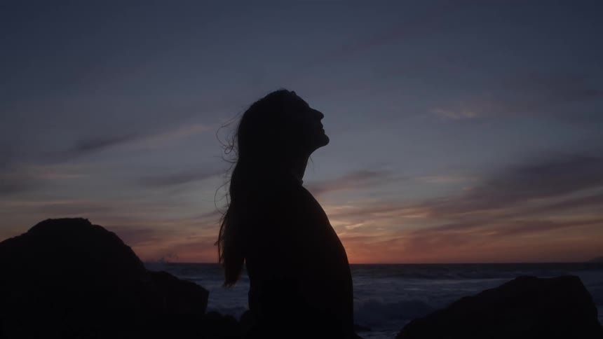A woman in silhouette tilts her head back beside the ocean at sunset hair lifting in the breeze with a calm reflective mood