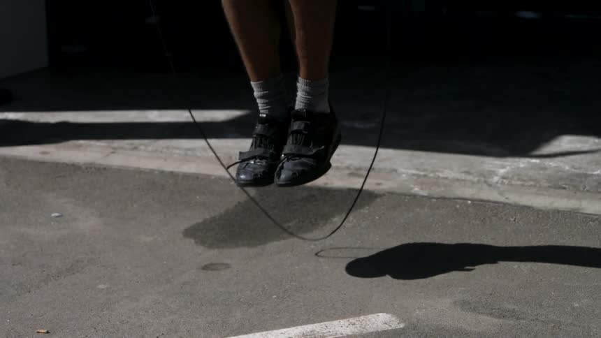 A person jumps rope with feet lifted above sunlit pavement as their shadow follows the arc of the rope