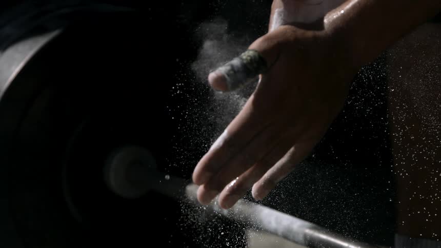 The video is an extreme closeup of a powerlifters chalkcovered hands as they prepare to lift a barbell the athlete has tape wrapped around their fingers and excess white chalk dust is visibly falling from their palms onto the bar below