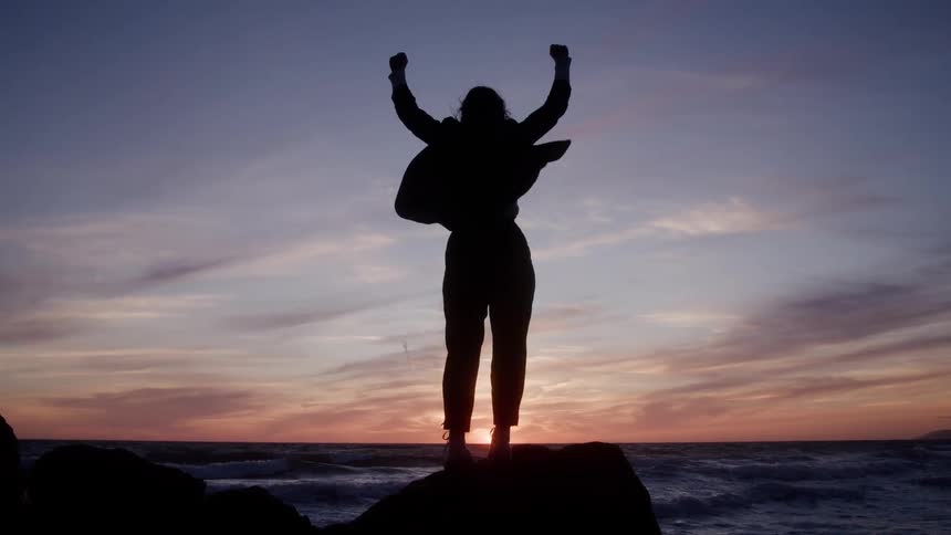 A woman on a cliff above the ocean raises her arms triumphantly