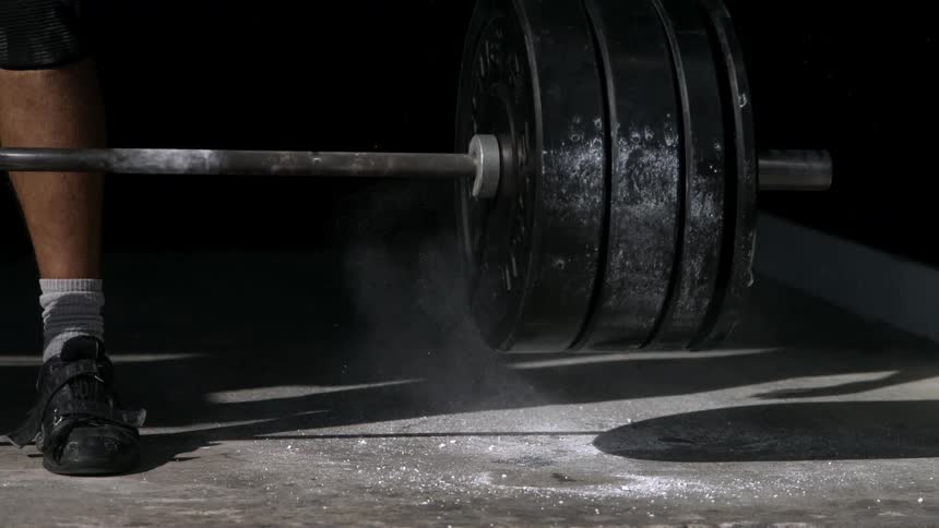The video is a lowangle closeup shot of a person setting down a heavily weighted barbell after a lift the scene is dark focusing on the barbell plates and the athletes feet in black shoes and white socks
