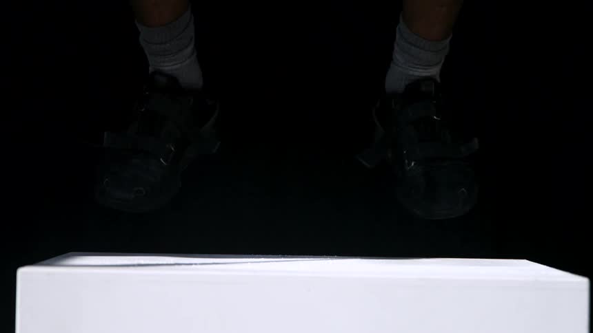 A low view shows the bare arms of a man and then his feet in black cycling shoes and grey socks landing on a white box kicking up a cloud of white powder against a completely dark background