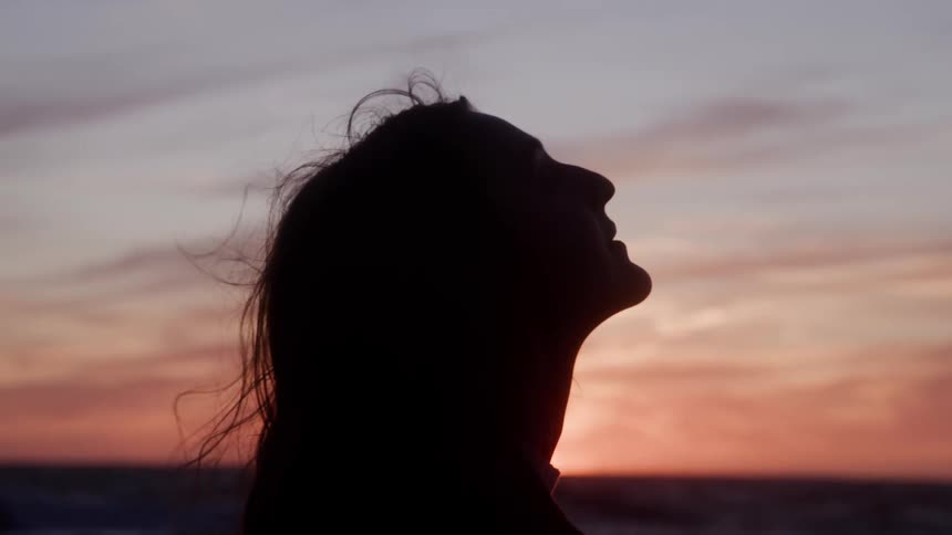 A silhouette profile view of a woman shows her head tilted back and face toward the sky as her hair gently moves in the breeze at sunset on the beach