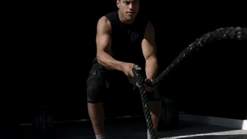 A strong man wearing a black tank top and knee brace is pulling thick black battle ropes out of a dark space working out intensely in the bright sunlight