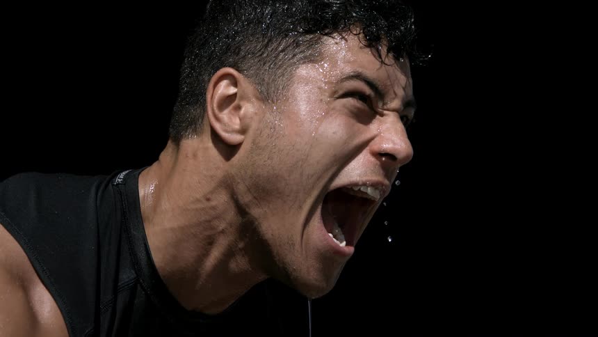 A young man expresses rage and anger with his mouth open as sweat drips down his face inside a dark room