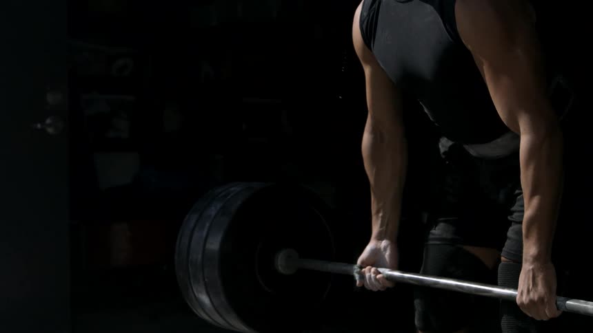 A muscular man wearing a black tank top and weightlifting belt deadlifts a heavy barbell with a focused expression in a dimly lit gym