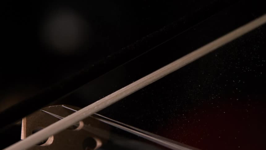 A close view shows a violin bow moving across the strings and wooden bridge of a dark string instrument scattering fine white rosin dust into the air