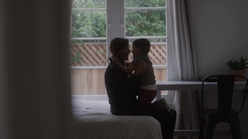 Woman sitting on a bed holds her son and rubs noses with the boy