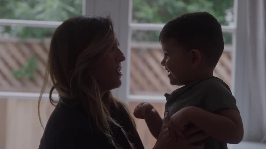 A woman holds a smiling young boy by a window as he playfully touches her nose