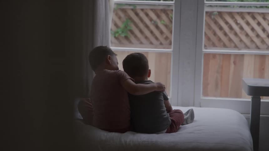 A pair of young boys sit side by side on a bed facing a bright window one with an arm around the other