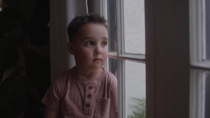 A young boy sits by a window gazing outside in soft daylight