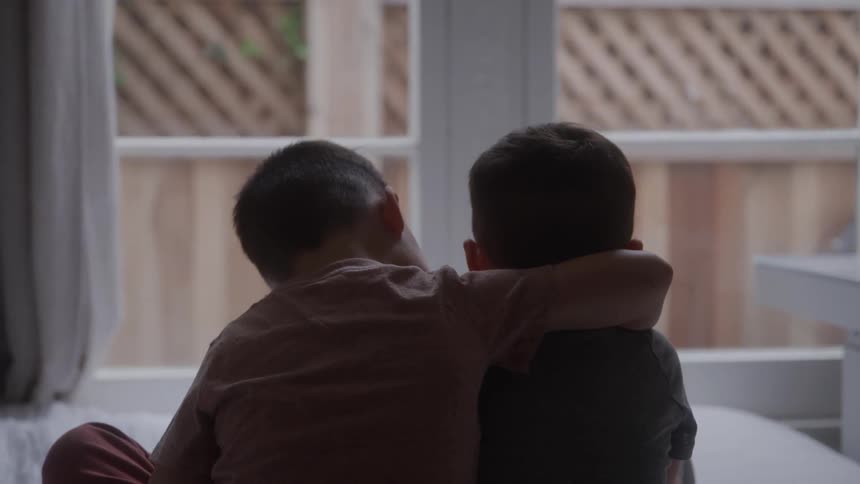 A pair of young boys sit by a window from behind one with an arm around the other in a quiet affectionate moment