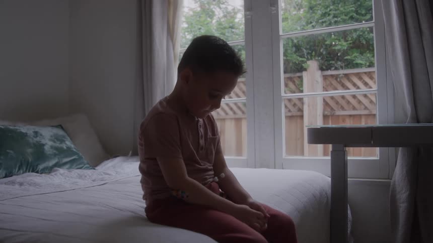 A child sits alone on a bed in his bedroom looking sad with his head down at midday