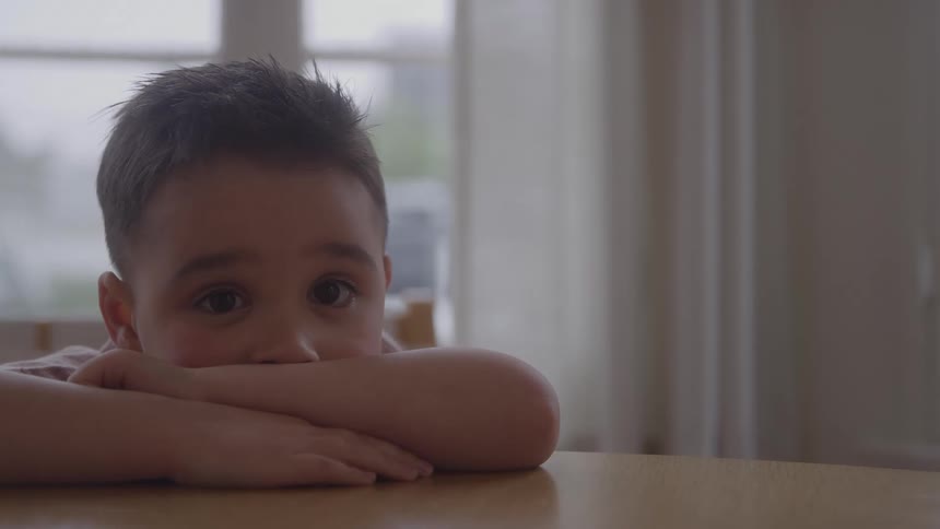 A close view shows a young boy with dark hair looking directly at the camera while resting his chin on his folded arms at a table indoors near a large bright window