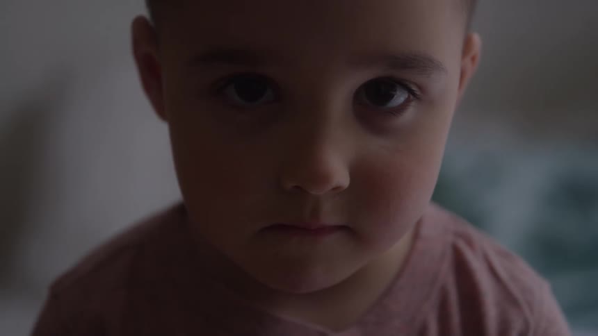 A young boys face staring forward the boy wears a pink garment the background is out of focus and dimly lit shadows obscure the facial features the scene is static and centered