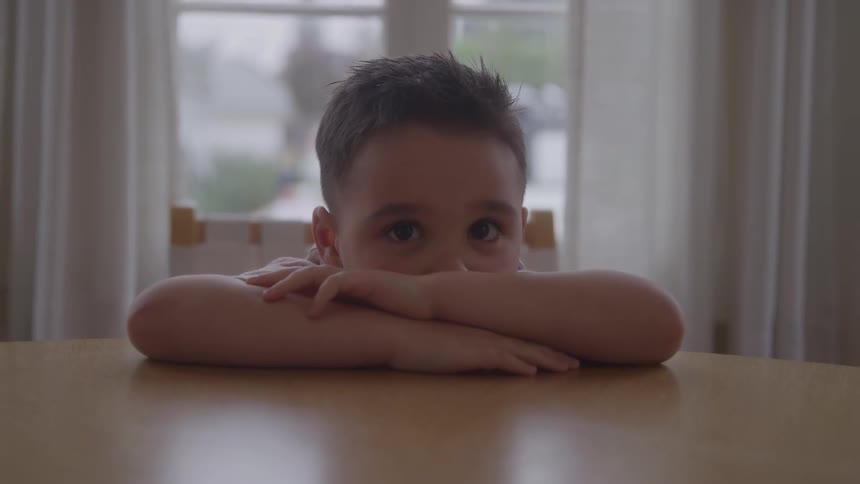 Young boy sitting at the table at home folds his arms in front of his face