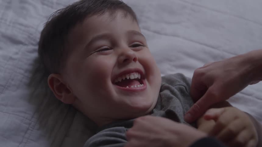 A young boy laughs joyfully while being tickled on a bed indoors