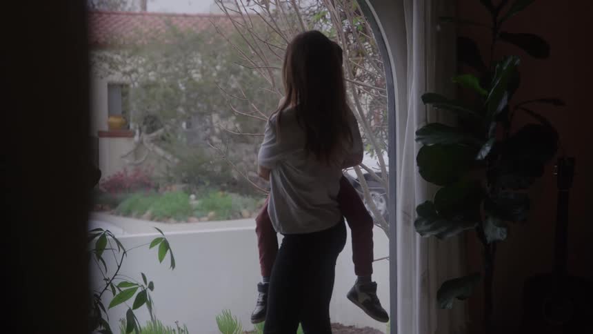 A woman holds her child as they look out a large window at home