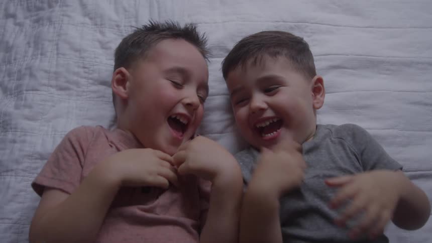 The video is an extreme closeup of two young children possibly brothers lying sidebyside on a bed or soft surface they are laughing heartily with wideopen mouths and appear to be enjoying a moment of playfulness