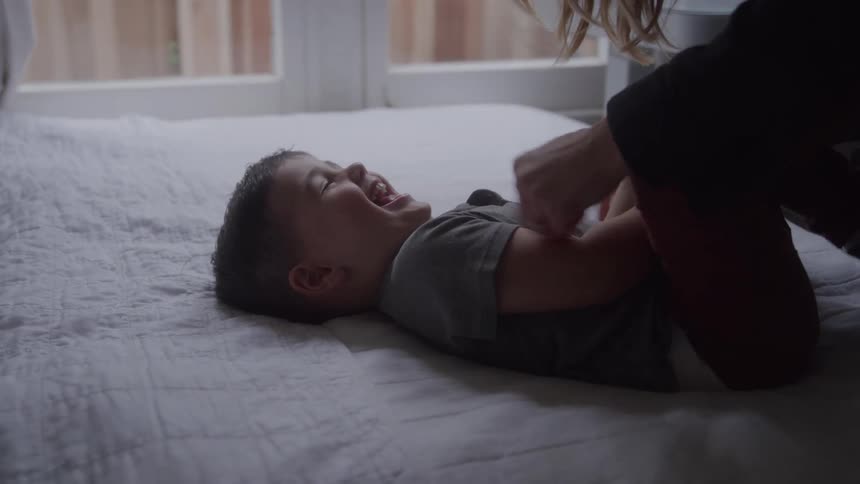 A mother tickles her laughing son on the bed in a bedroom