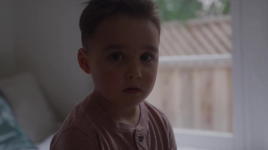 A closeup shot focuses on a young boy whose face shifts from looking pensive to one of slight surprise or curiosity the lighting is dim suggesting he is standing near a window that offers a view of a fence outside