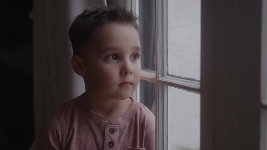 A young boy with a serious expression wears a pink button up shirt while looking out a window on a cloudy day with soft light illuminating his face