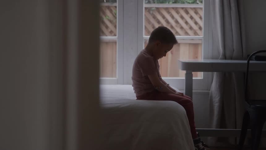 The video shows a young child in a pink shirt sitting alone on the edge of a bed in a dimly lit room the child is facing the window their head bowed creating a somber introspective silhouette against the natural light