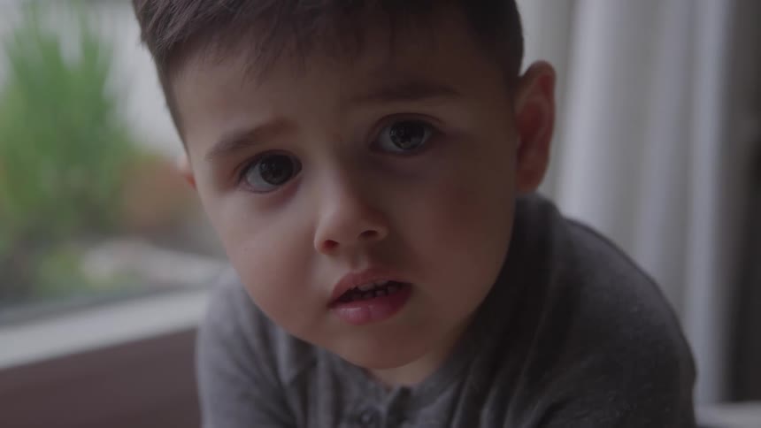 The video is a tight closeup focusing on the face of a young boy with wide expressive eyes he is wearing a grey buttonup shirt and is looking directly at the camera with a look of concern or gentle inquiry