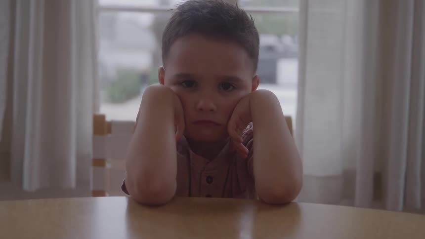 A young boy rests his chin on his hands while sitting quietly at a table indoors
