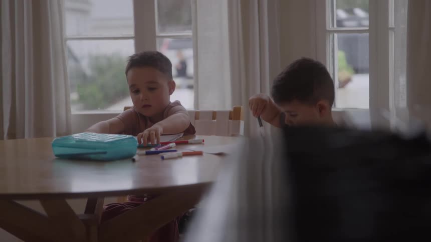 Two young boys sit at a wooden table in a well lit room coloring with markers and paper with windows in the background showing a cloudy day