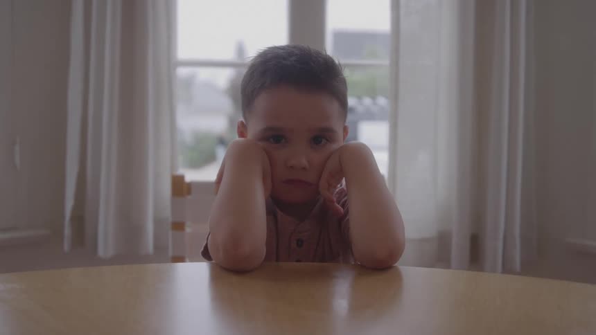 A close view shows a young boy with a serious expression and a pink shirt resting his head in his hands against a light colored background