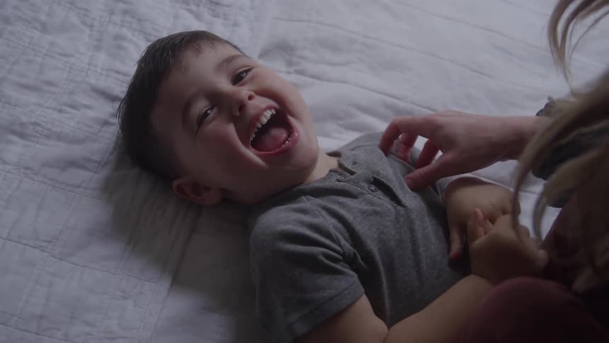 A child lies on the bed laughing as his mother tickles him filling the room with playful joy