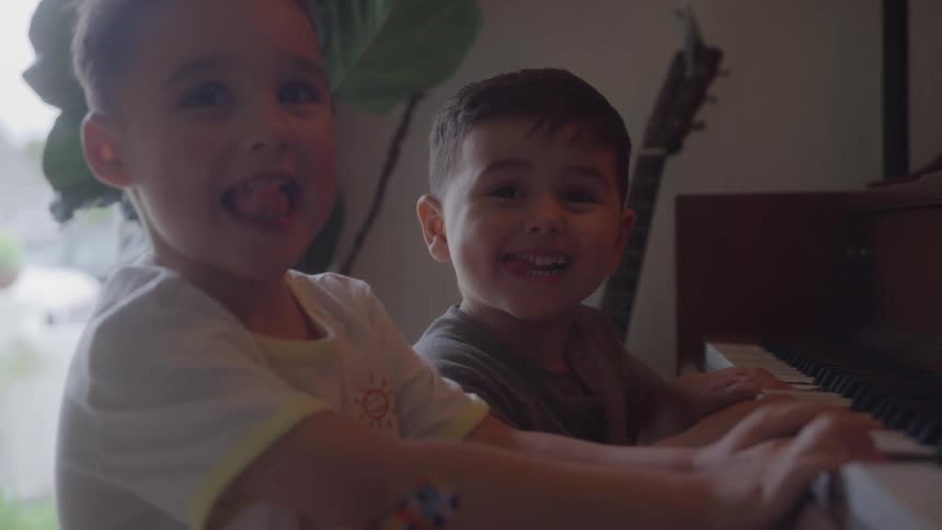 Two smiling young boys play the keys of a dark upright piano together making silly faces with their tongues out in a dimly lit room with a plant and guitar visible