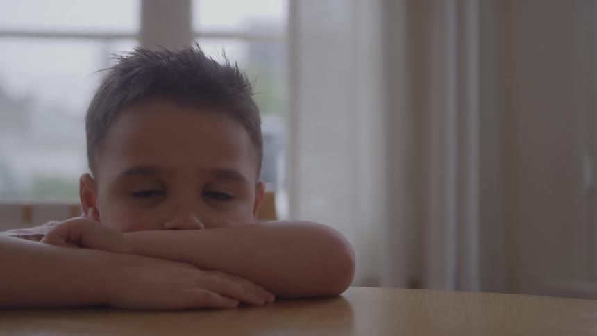 A young boy with messy dark hair peeks over his crossed arms resting on a wooden table with a window and soft daylight in the background