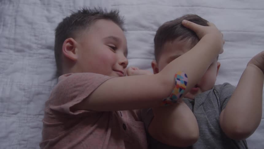 Two young boys are lying sidebyside on a white quilted blanket the boy on the left reaches out his arm toward the other boys face the boy on the right reacts by raising his hands to his head and opening his mouth both boys wear shortsleeved shirts