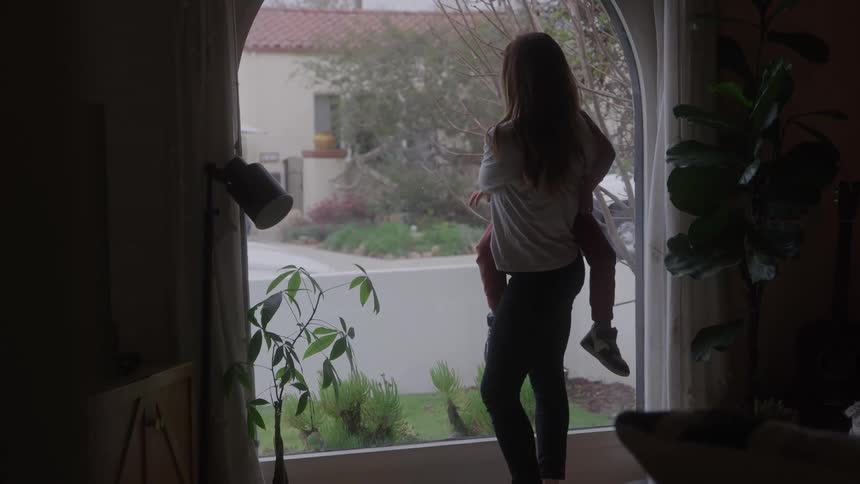 The video is an interior shot of a woman holding a young child up to a large window they are looking outside at a rainy or overcast day with a residential street visible in the background