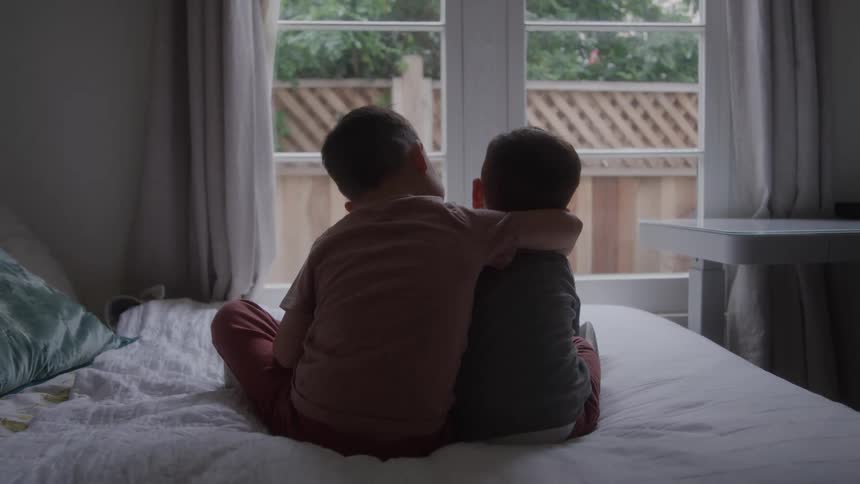 The video shows a comforting scene of two young children possibly brothers sitting with their arms around each other on a bed they are facing away from the camera toward a window with diffused light giving the scene a warm quiet and tender quality
