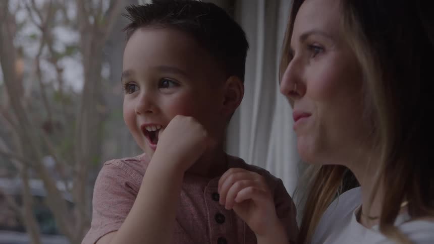 A mother and son smile together while looking out a window at home