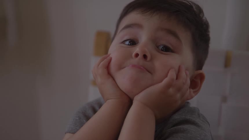 A closeup of a young boy looking up at the camera the boy rests his chin on his cupped hands he wears a gray shirt the background is out of focus with light colors the lighting is soft and ambient