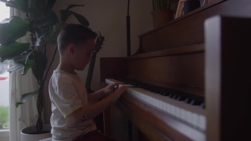 The video features a young boy sitting at an upright piano near a window attentively playing the keys the scene is dimly lit with natural light coming from the side emphasizing the childs focus on the musical performance