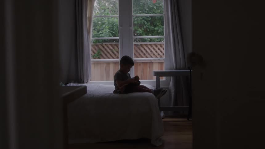 A young boy sits on a bed holding a toy near a window in a dimly lit bedroom