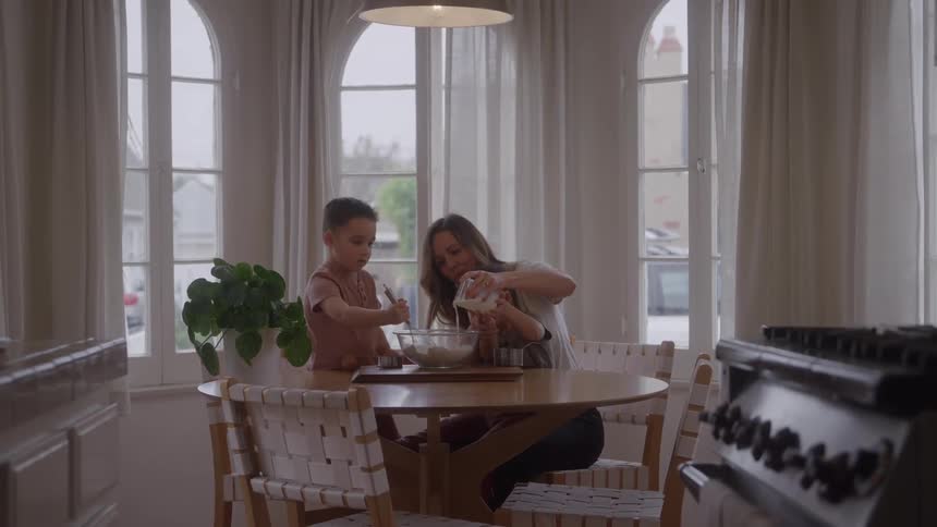 A smiling woman and a young boy mix ingredients in a bowl on a round wooden table near large arched windows with sheer curtains in a brightly lit kitchen