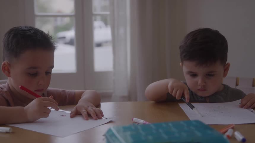 Two young boys sit at a table drawing with markers on paper indoors