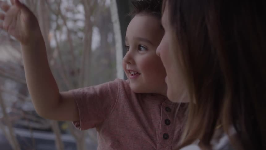 A mother and son smile as they look out a window together inside their home