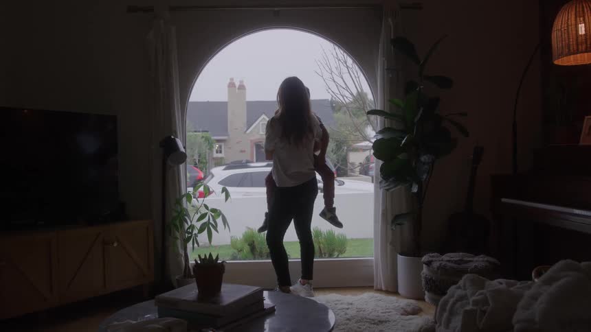 A mother holds her young son while standing at a large window at home