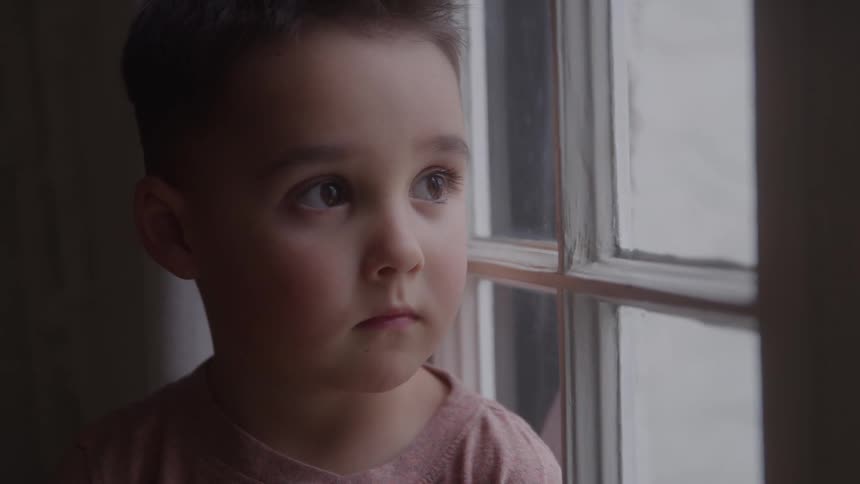 The video is a closeup on a young boy who is looking intently out of a window the soft overcast light coming from the window creates a pensive and slightly somber mood around his face