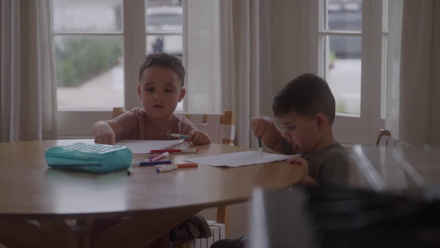 A pair of young boys sit at a round table by bright windows one sorting colorful markers while the other concentrates on his drawing